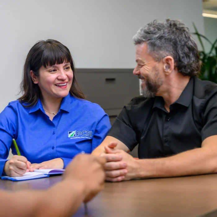 A young woman on the left wearing a PCSI shirt and a middle-aged man chatting on the right. He is wearing hearing aids and she is taking notes in a notebook.